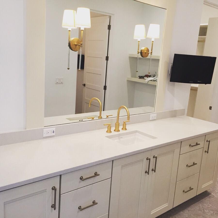 Bathroom vanity with large mirror, gold fixtures, white cabinets, and a wall-mounted TV.