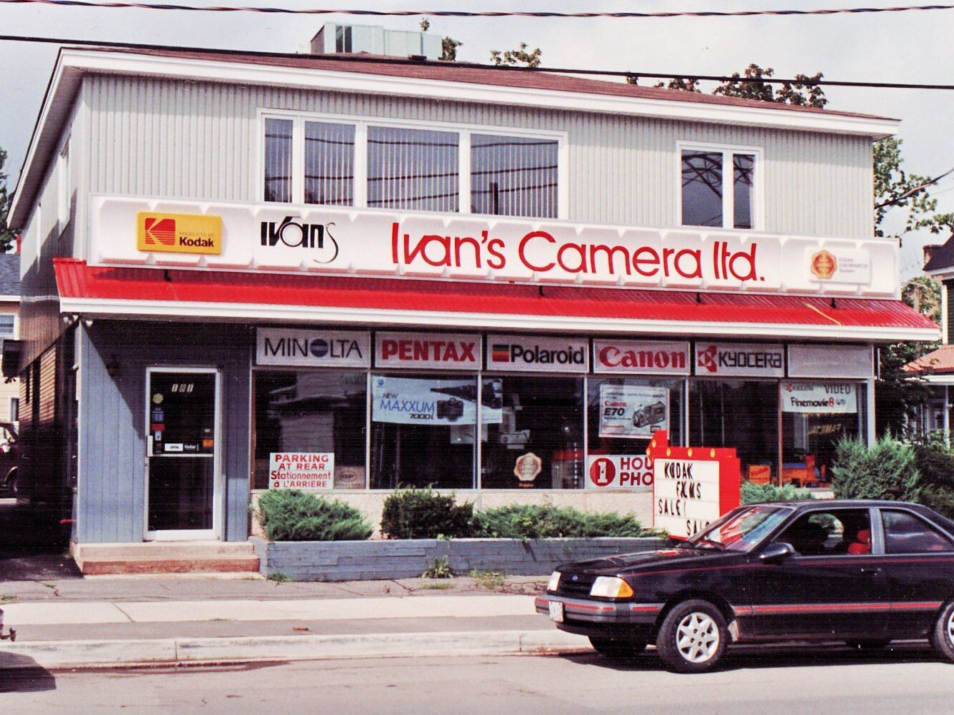 Storefront of Ivan’s Camera Ltd. with film brand signs and a car passing on the street