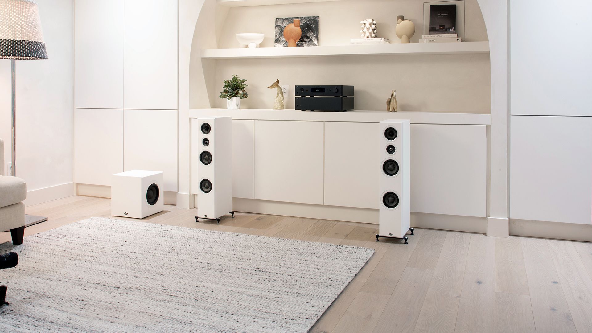 Bright minimalist living room with white speaker towers under a shelf and a light rug on the floor