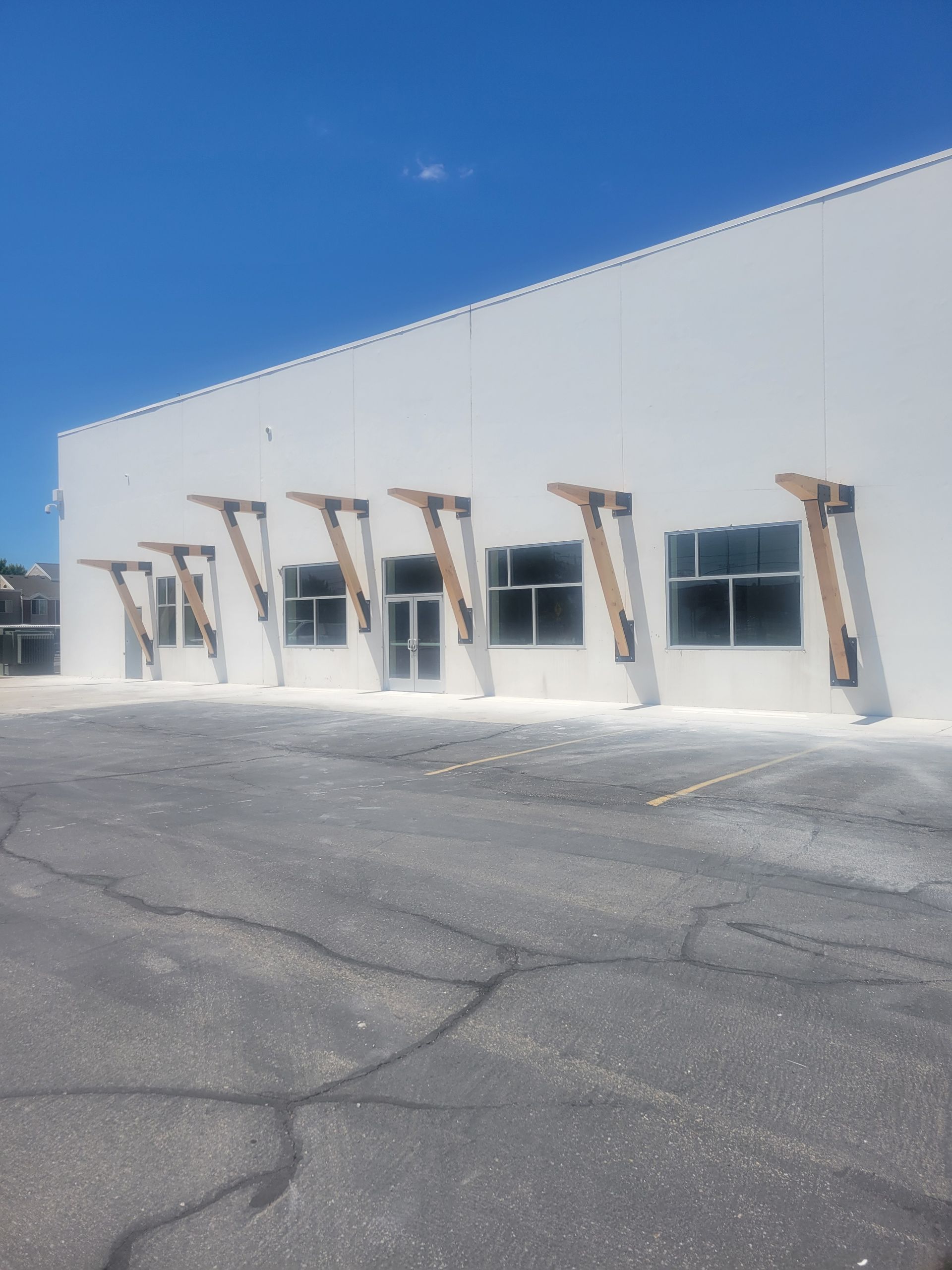 White industrial building with windows and wooden awnings beside an empty asphalt lot under a blue sky