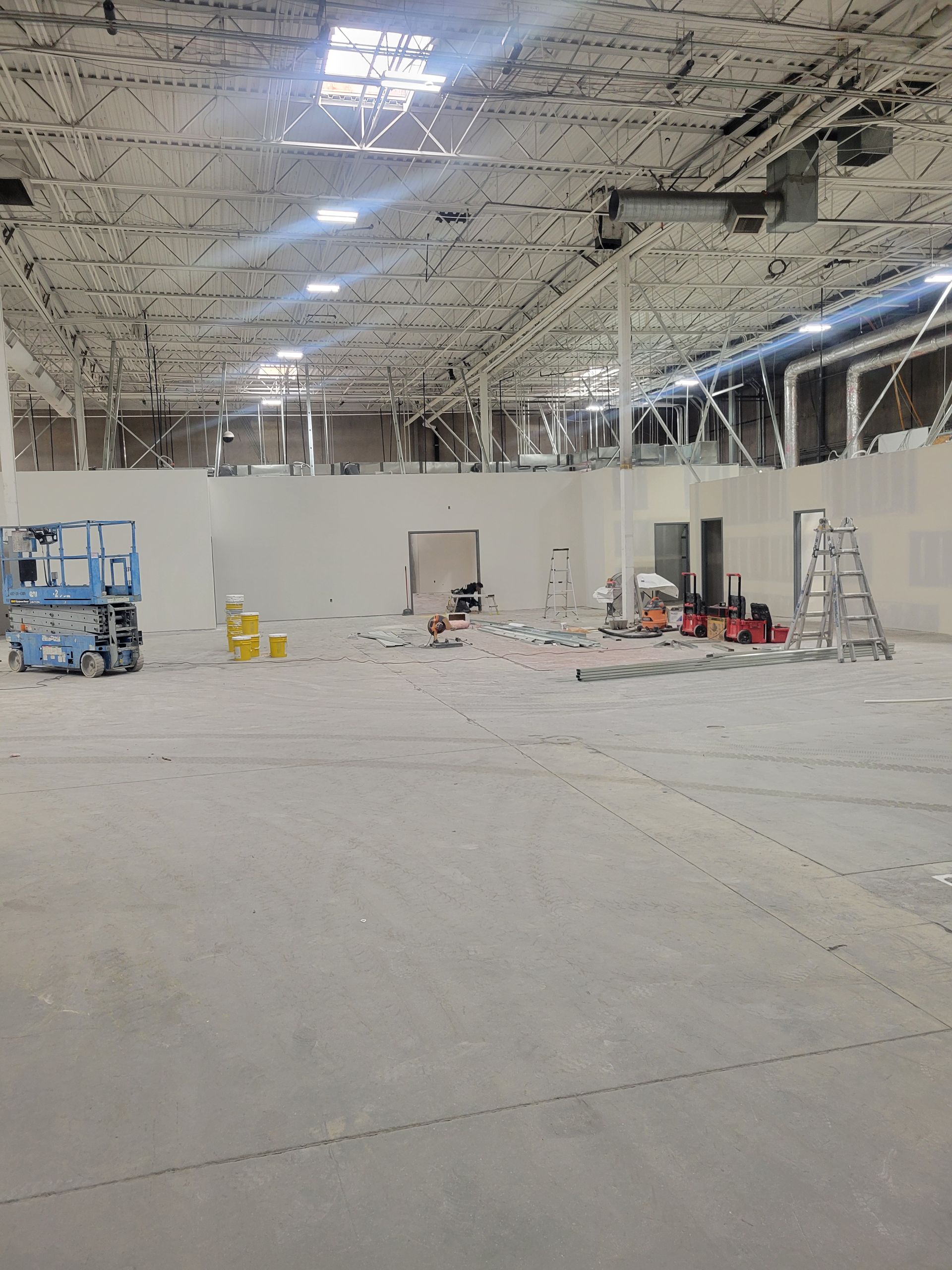 Large indoor construction site with unfinished ceiling, white walls, ladders, and equipment scattered across the floor