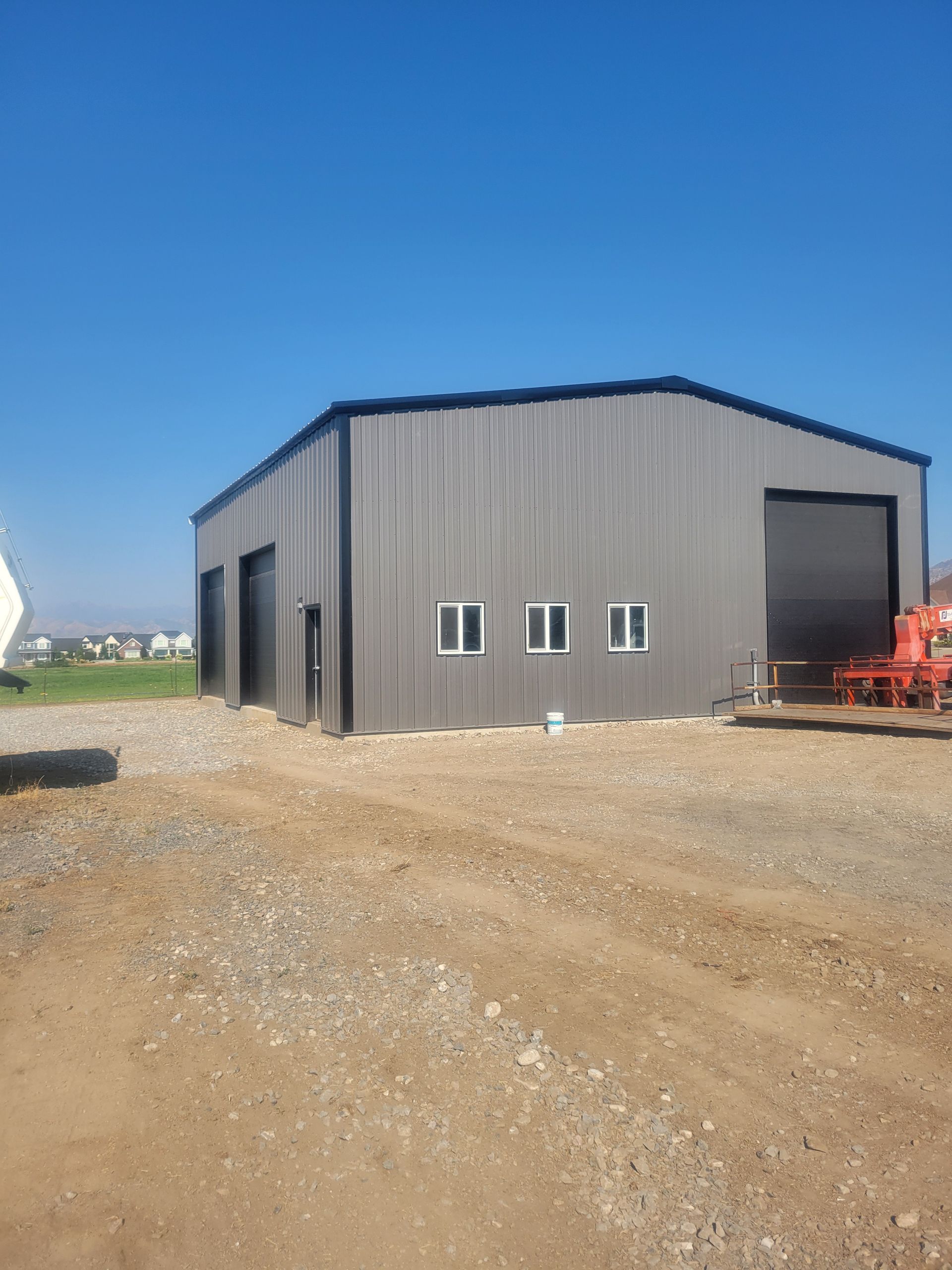 Gray metal warehouse building on a gravel lot under a clear blue sky, with red equipment by the open door