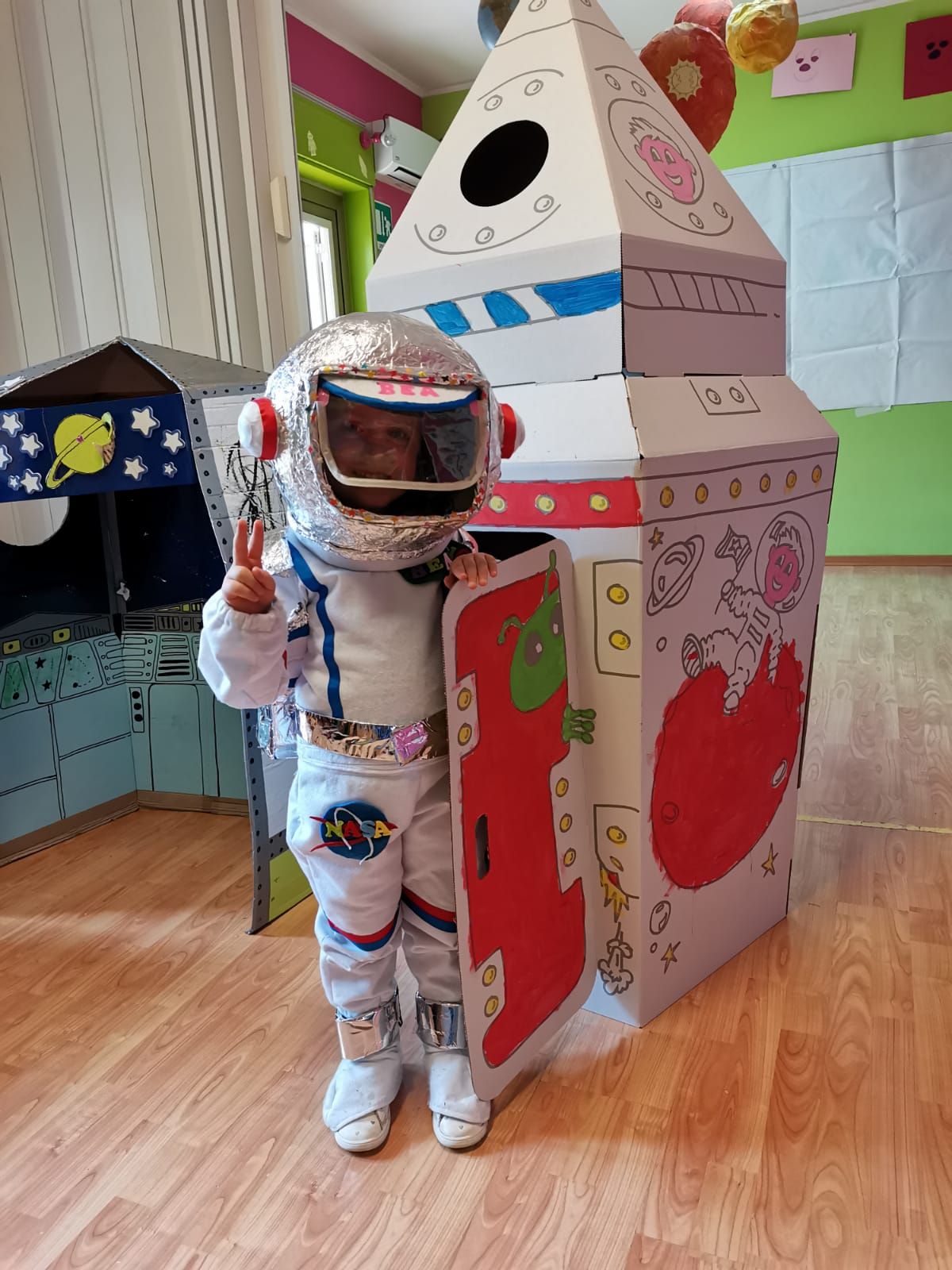 Un bambino vestito con un costume da astronauta bianco è in piedi accanto a un razzo di cartone colorato in un'aula scolastica.