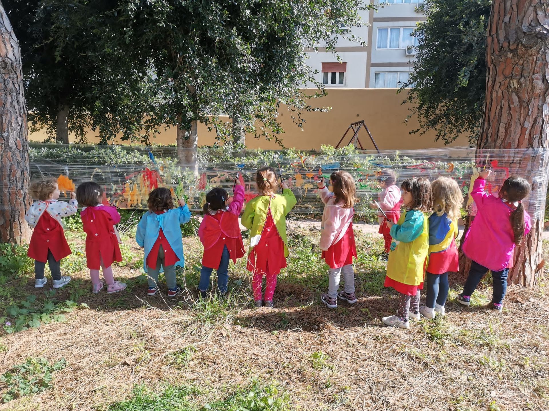 Bambini con cappotti colorati in piedi all'aperto in un giardino, di fronte a un'aiuola e agli alberi.