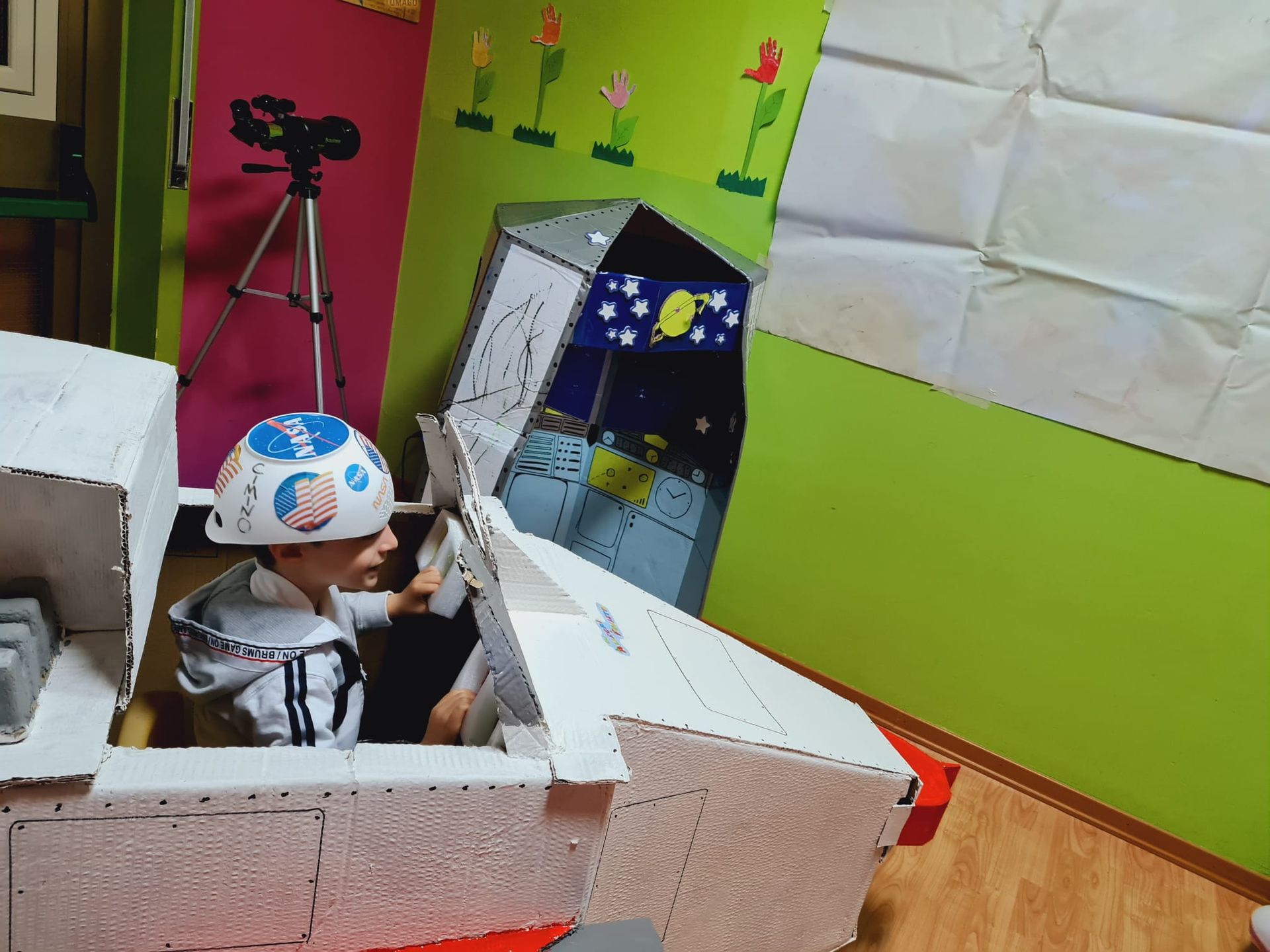 Un bambino con il casco è seduto in un'auto di cartone in una colorata stanza dei giochi, con un treppiede per la macchina fotografica nelle vicinanze.