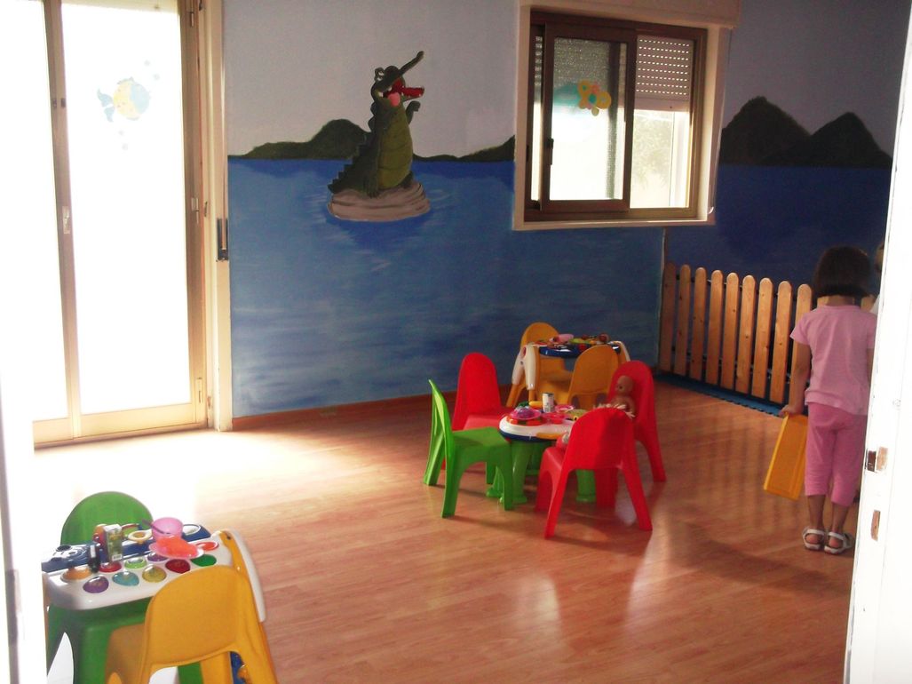 Sala giochi per bambini con murale raffigurante un coccodrillo, tavolini, sedie.