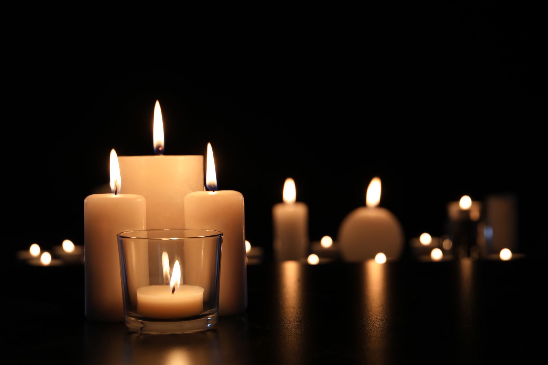 A group of white candles with glowing flames sits on a dark surface, with soft bokeh lights in the background.