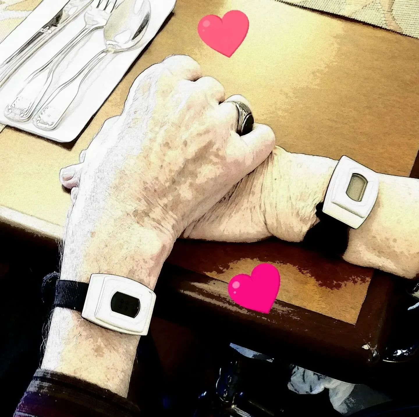 Two hands, wearing digital health-monitoring watches, are clasped together on a table with silverware. Pink hearts overlay.