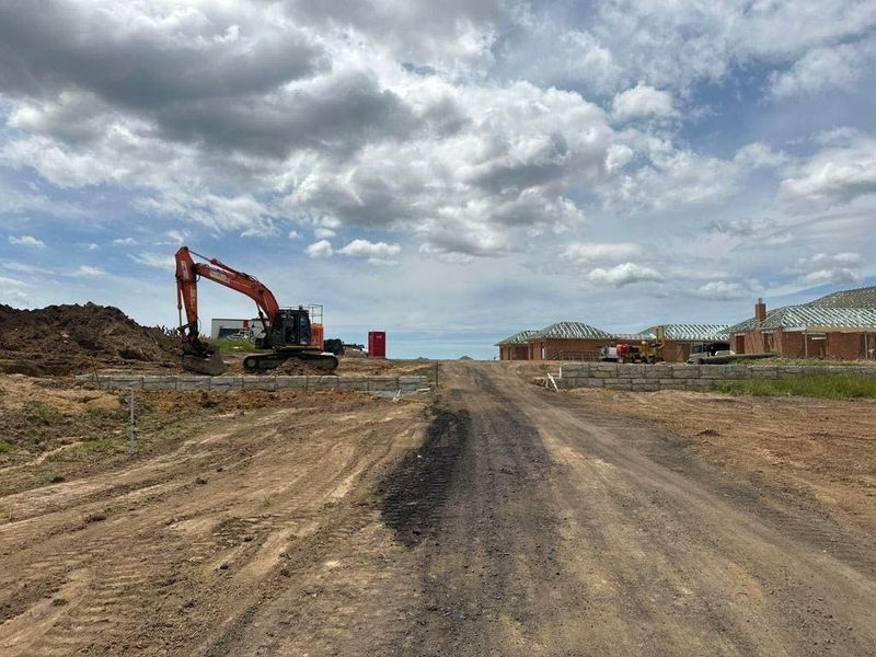 An Excavator Is Driving Down A Dirt Road Next To A House — About Time Plumbing & Civil Construction in Moss Vale, NSW