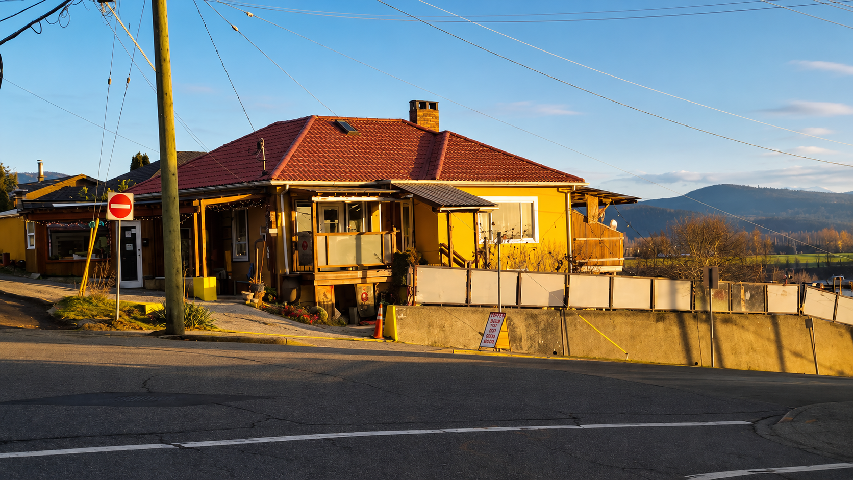 Yellow house with red roof and porch beside a road, with mountains in the background.