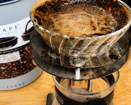 Coffee grounds in a metal pour-over dripper beside a coffee bean bag and glass carafe