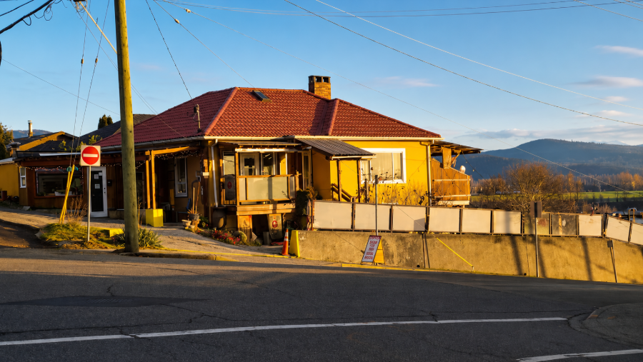 Yellow house with red roof beside a road, fenced yard, and mountains in the background at sunset