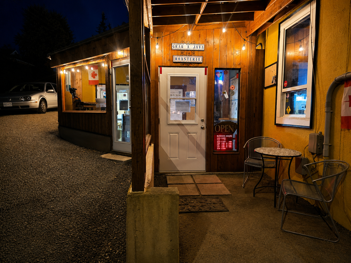 Small wooden storefront at night with warm lights, gravel path, and a bench by the entrance.