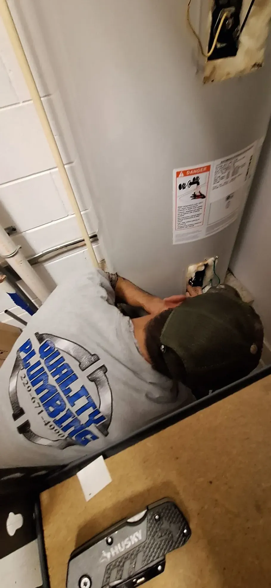 A plumber works on a water heater. He is wearing a Quality Plumbing t-shirt and a green cap. A Husky knife is nearby.