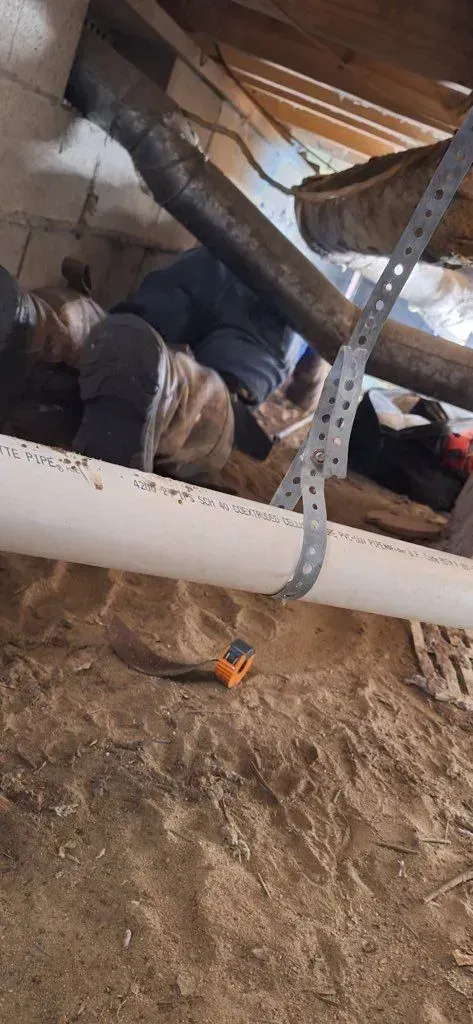 Person working under a building, possibly on plumbing. White pipe visible, with tool on the ground.