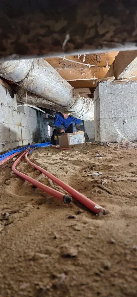 Red pipes in a crawl space, near concrete block walls and wooden beams, on sandy soil.