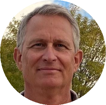 Portrait of a man with short gray hair, smiling slightly outdoors with trees and blue sky behind him