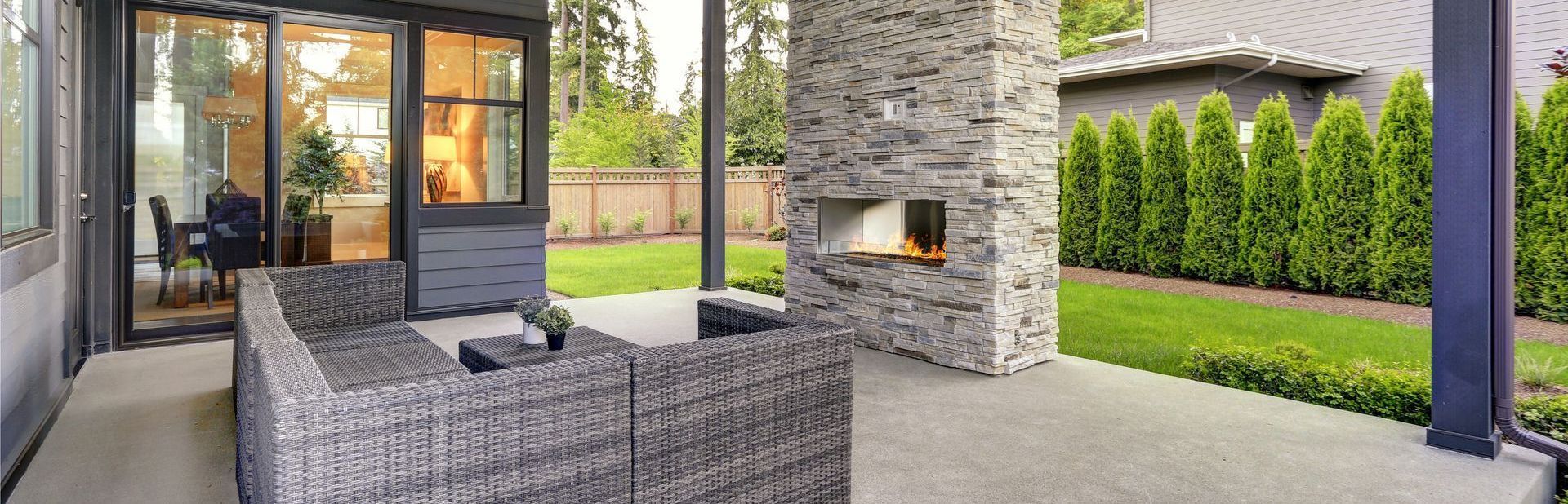 Covered patio with gray wicker seating, stone fireplace, and green backyard hedge