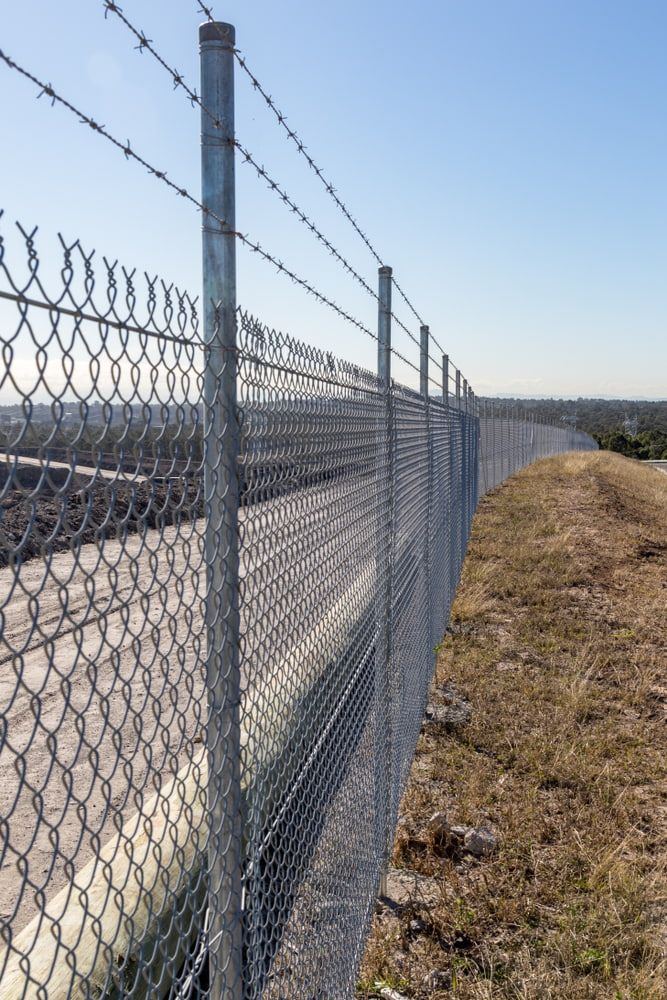 A Chain Link Fence With Barbed Wire Along the Side of a Road — AM & JD Contracting In Muswellbrook, NSW