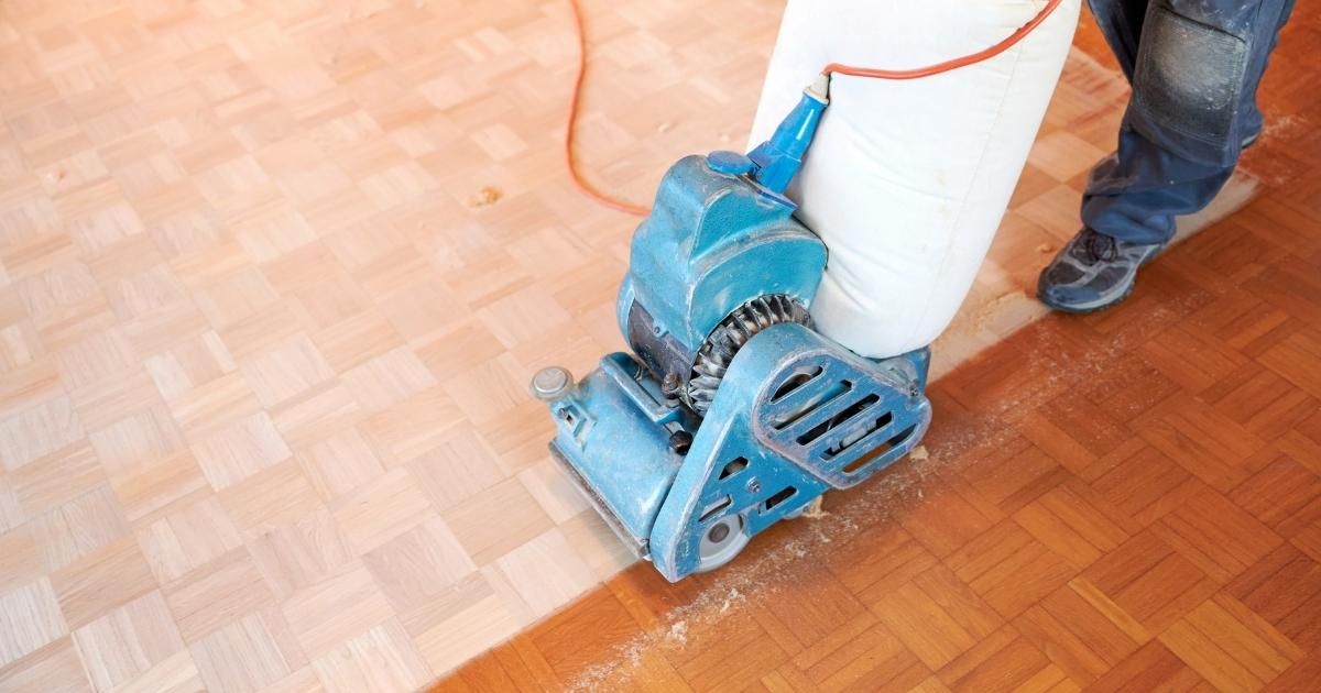 Blue floor sander polishing a wooden parquet floor, with a person standing beside it