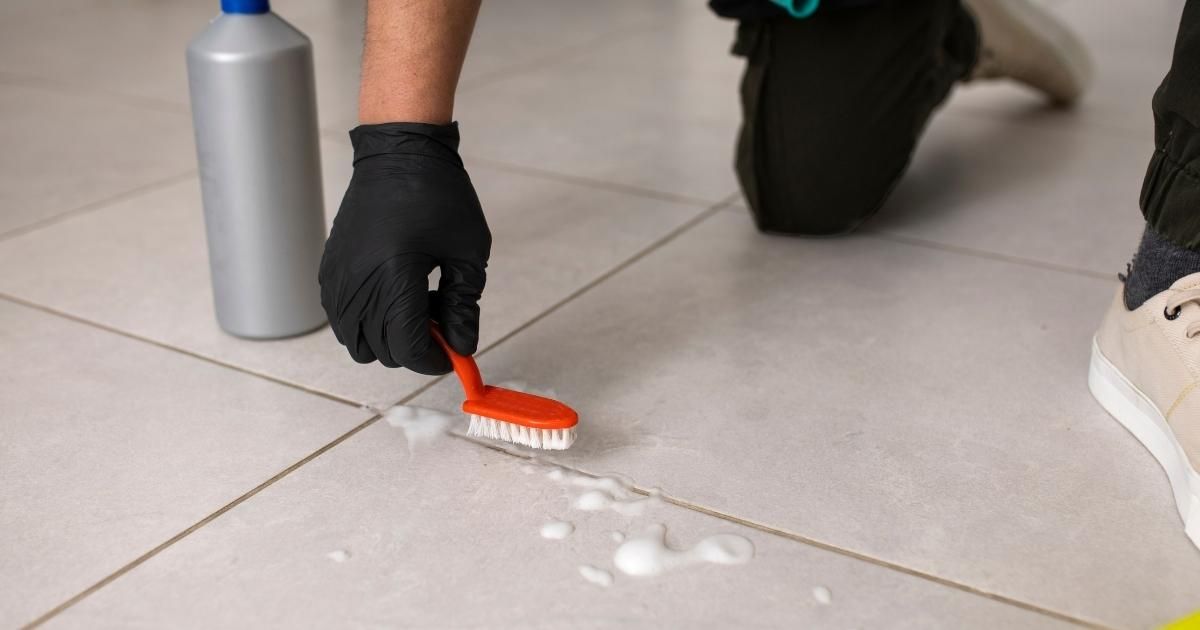 Gloved hand scrubbing white foam from a tiled floor with an orange brush beside a cleaner bottle