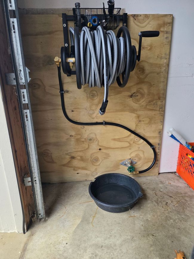 A wall-mounted hose reel with a black hose connected to a water spigot above a black drip pan on a garage floor.