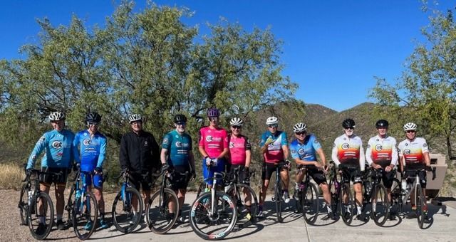 Group of cyclists posing with bikes outdoors, sunny day. Trees and mountains in background.