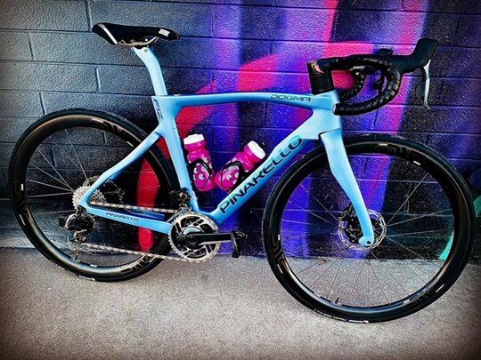 Light blue Pinarello road bike with black components, against a graffiti wall, two water bottles.