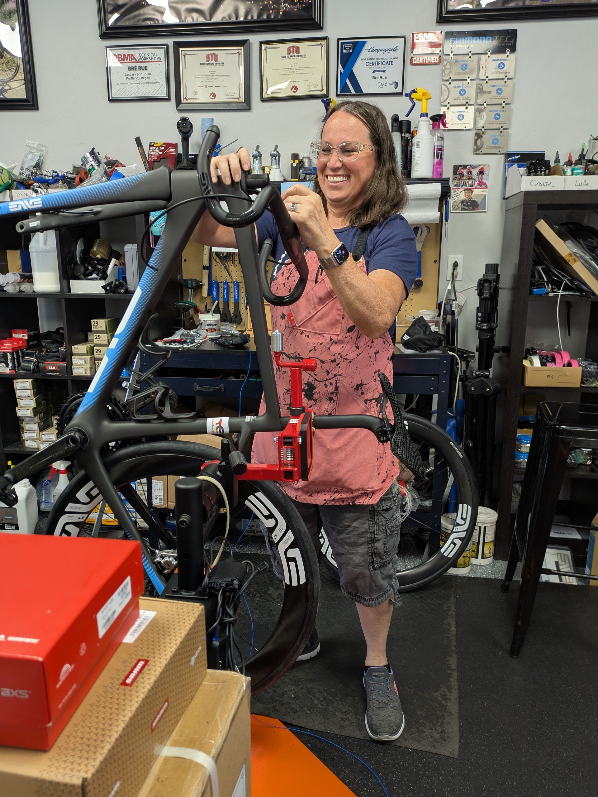 Man holding a bicycle wheel in a bike shop. Red and orange frame, black tires, tools, and posters visible.