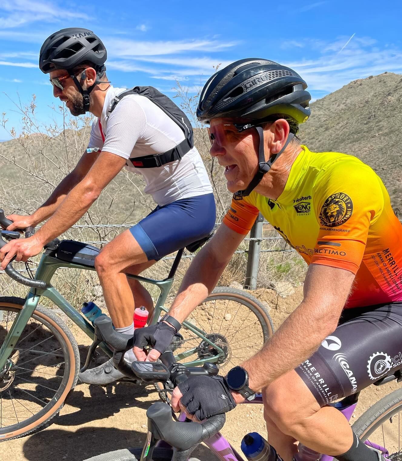 Two cyclists on bikes outdoors on a dirt trail; one in a yellow-orange jersey, the other in white and blue.