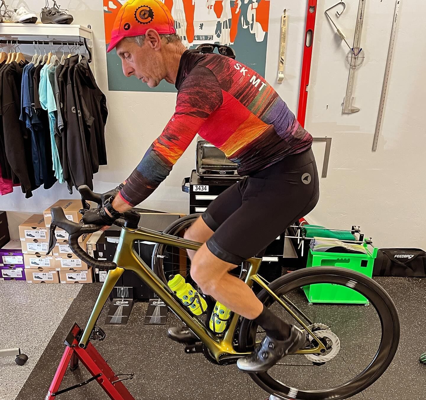 Man on stationary bike, wearing cycling gear. Indoors, yellow bike, black shorts, colorful top, training.