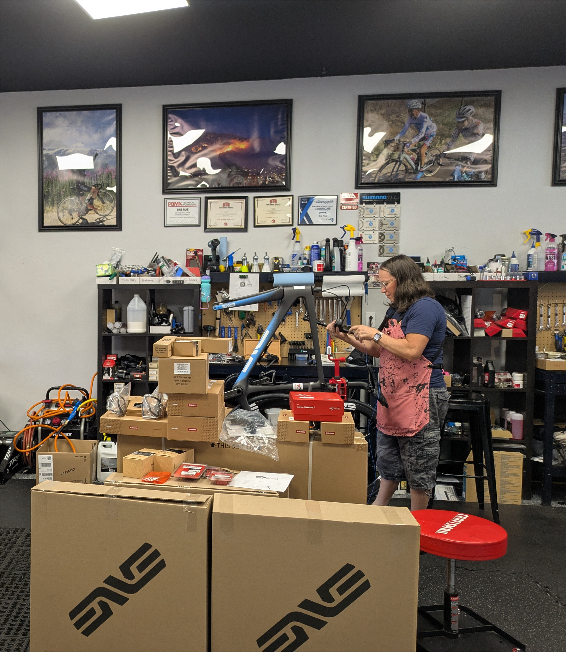 Two people in a bike shop. One repairs a bike, the other organizes parts. Bikes and supplies surround them.