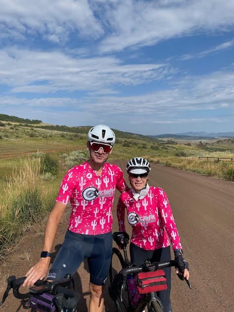 Two cyclists in pink jerseys smiling beside a dirt road in a wide, open landscape under a cloudy sky