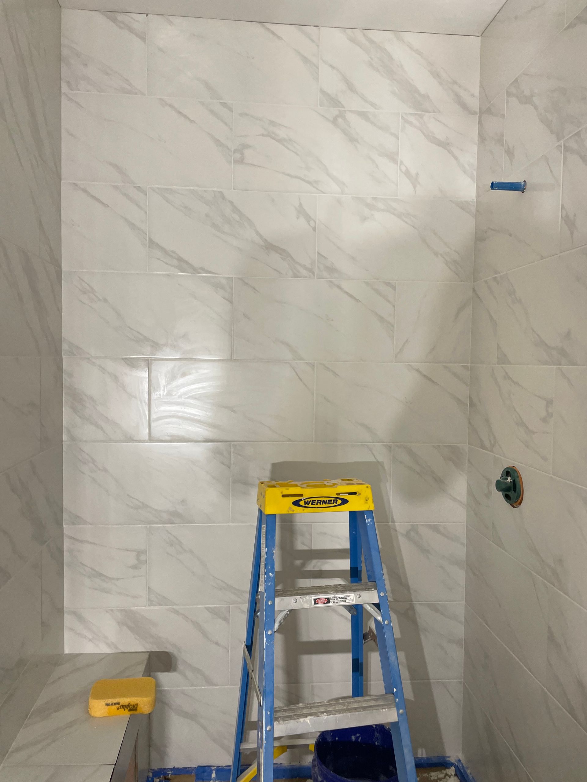 Bathroom wall with large marble-look tiles, a blue ladder, and a tile cutter on the floor.