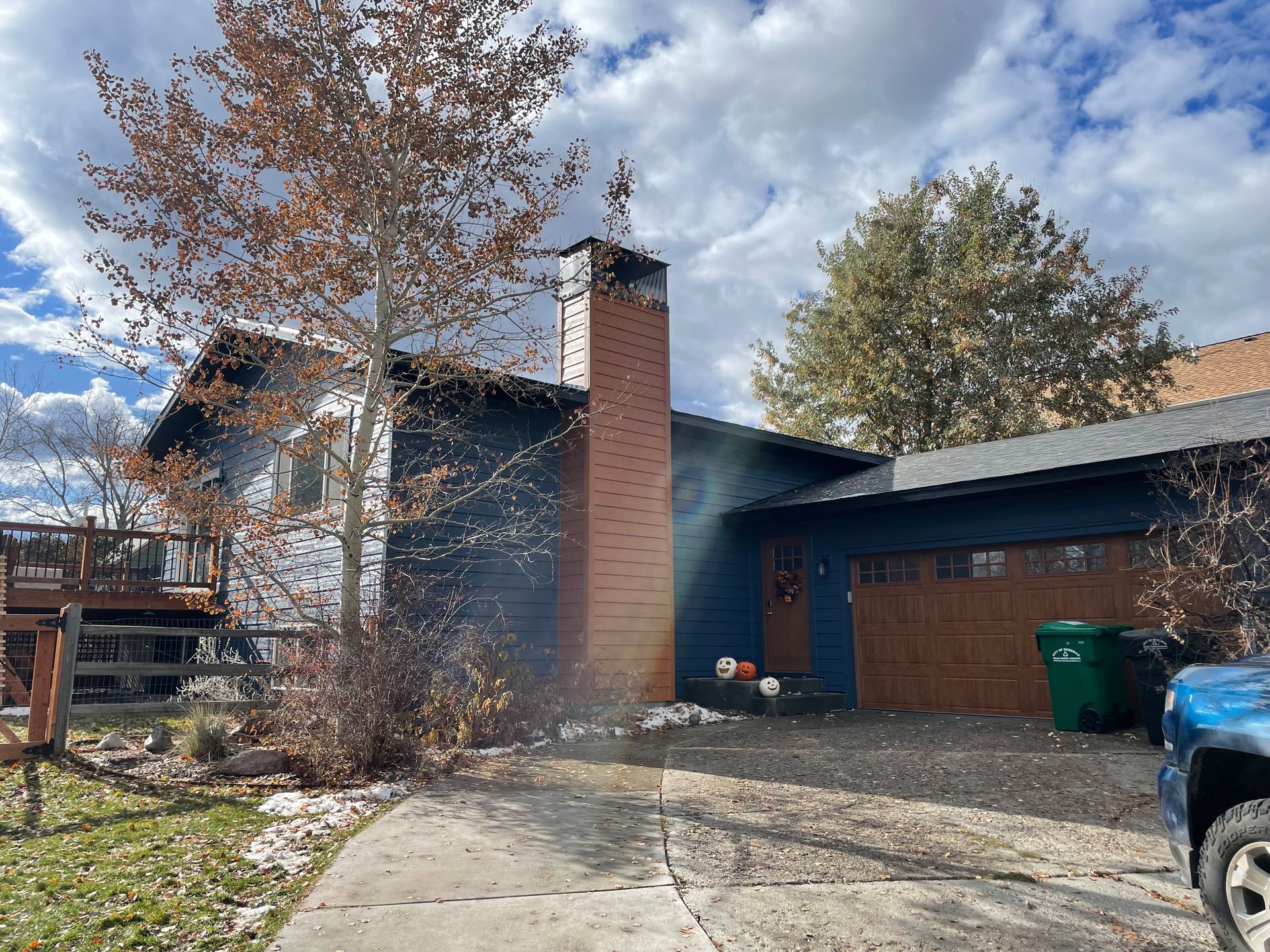 Suburban house exterior with a chimney, autumn trees, driveway, and parked truck under a cloudy sky