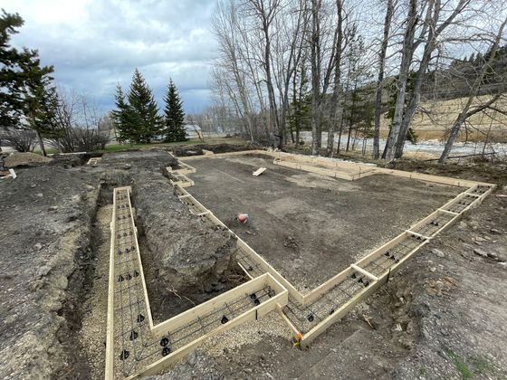 Wooden foundation framing in a dirt yard, with bare trees and cloudy sky in the background