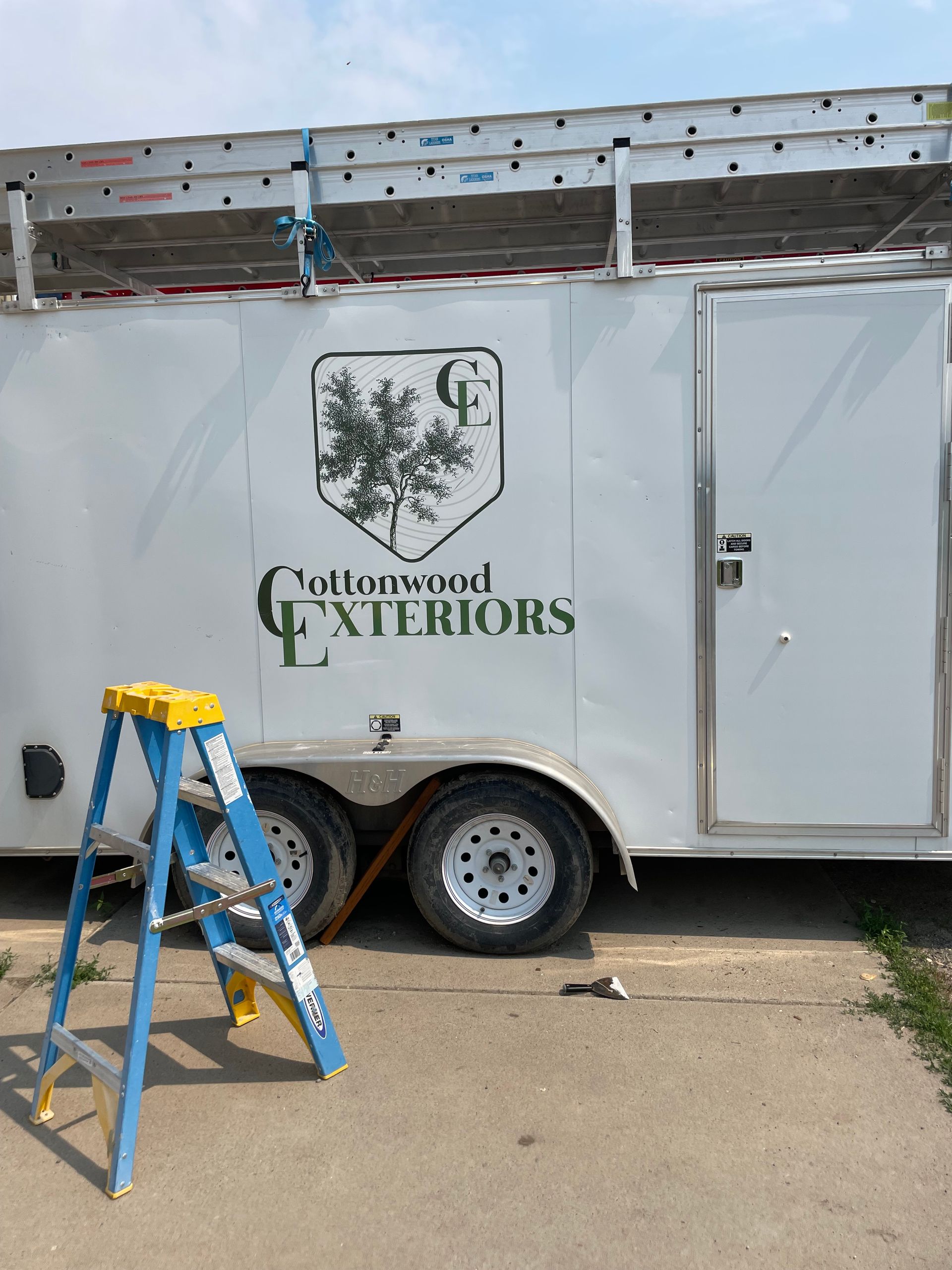 Blue ladder beside white GJ Exteriors trailer with roof work materials at a job site