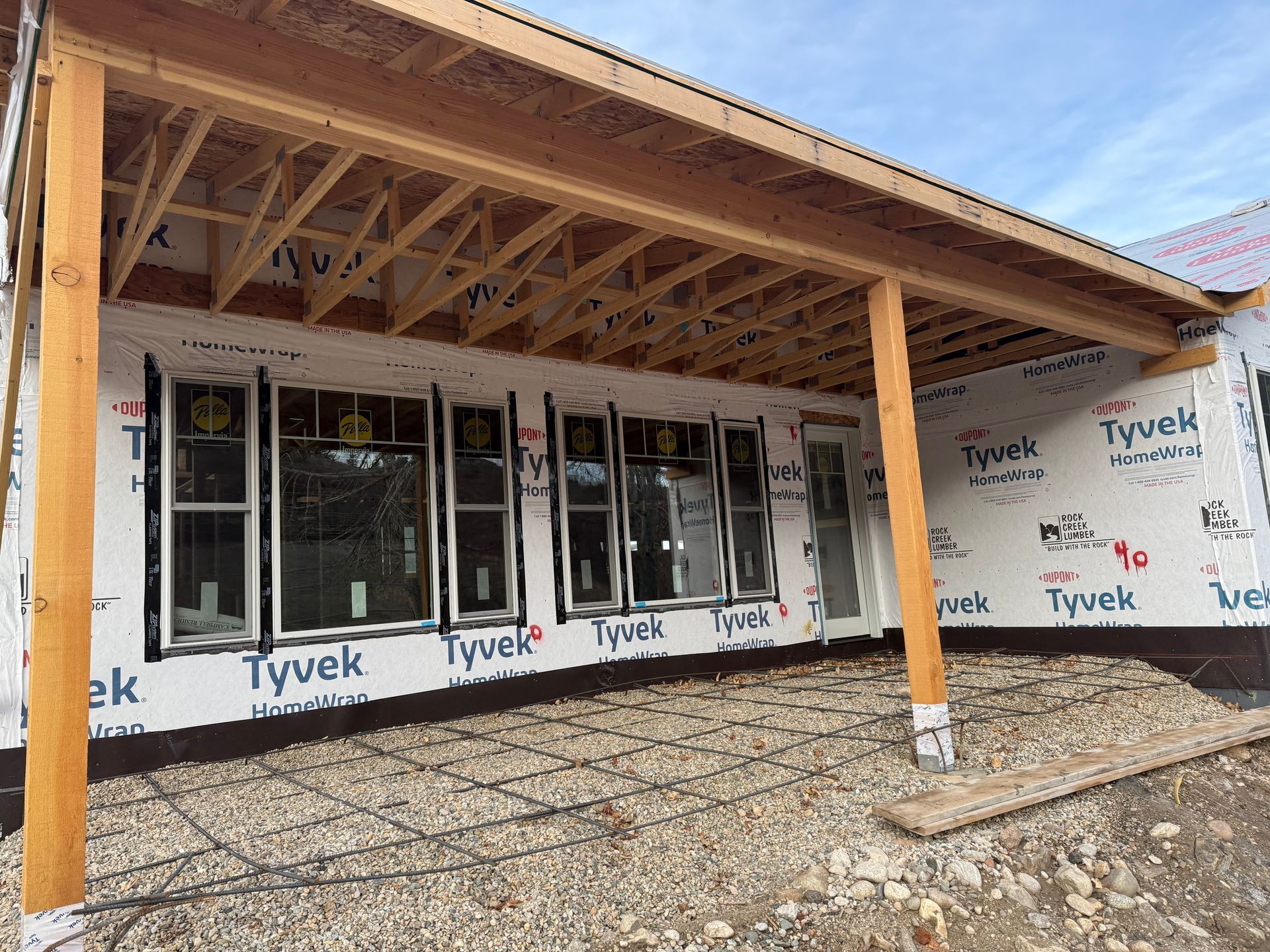 Partially built house exterior with exposed wood framing, Tyvek wrap, and gravel ground under a covered roof.