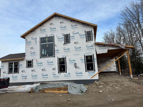 Unfinished house wrapped in white housewrap, with exposed framing and a covered porch on a dirt lot.