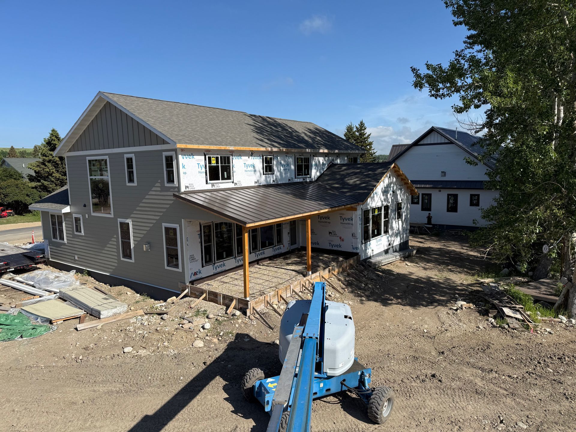 Partially built two-story house with wooden porch framing and a blue lift in front on a dirt lot