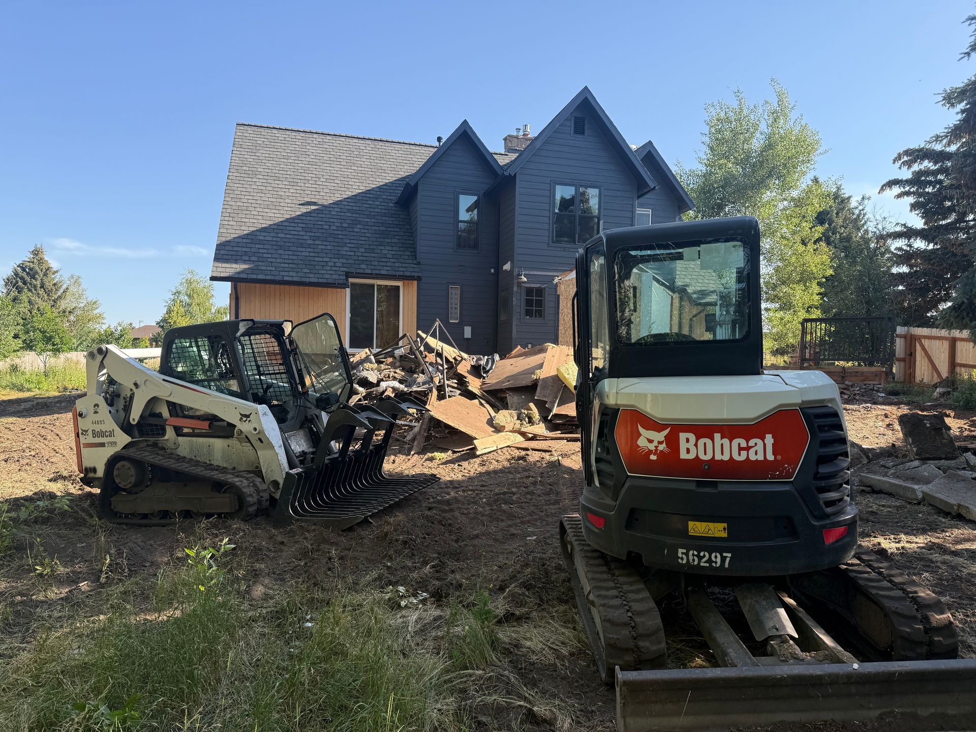 Two construction machines clearing debris in front of a dark blue house under a clear sky