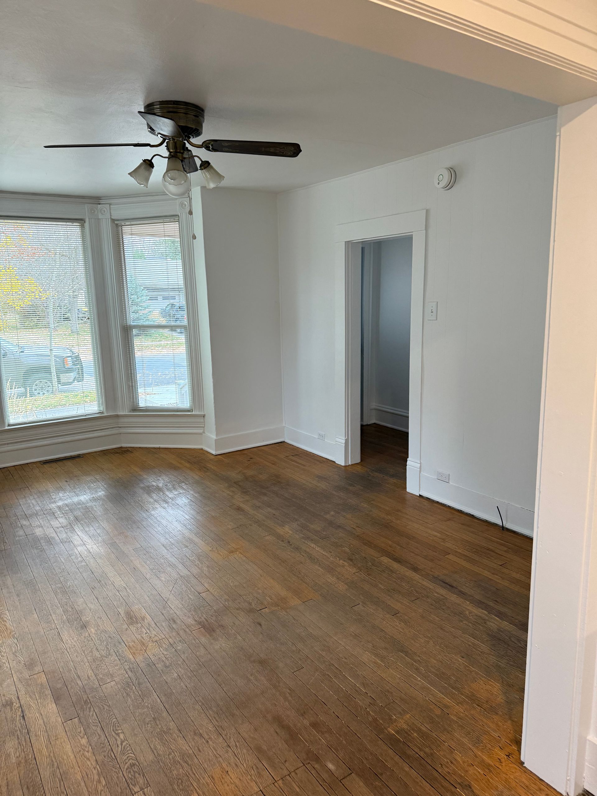 Empty room with wood floors, white walls, ceiling fan, and large windows with shutters