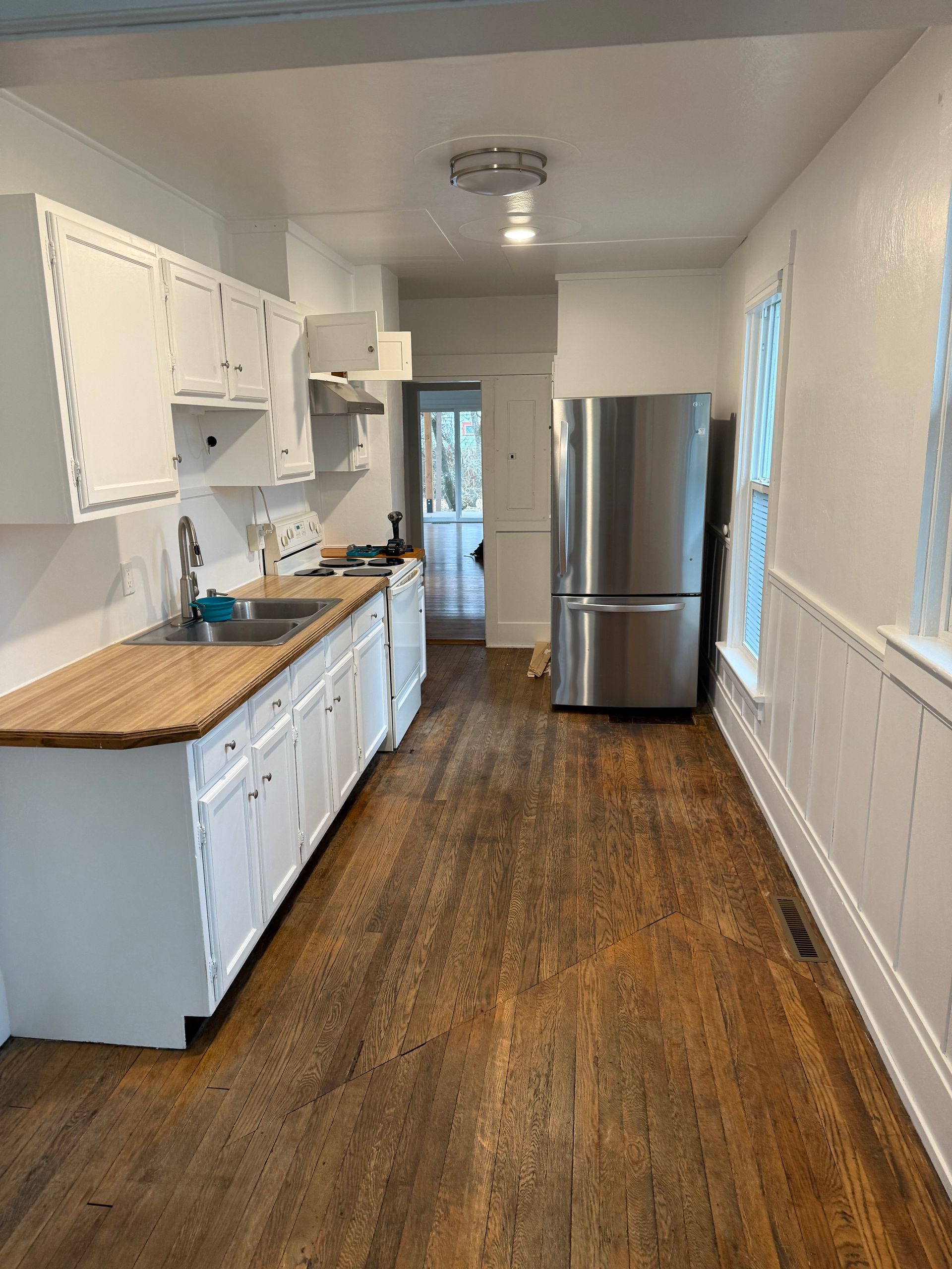Narrow galley kitchen with white cabinets, wooden counters, and a stainless steel refrigerator.