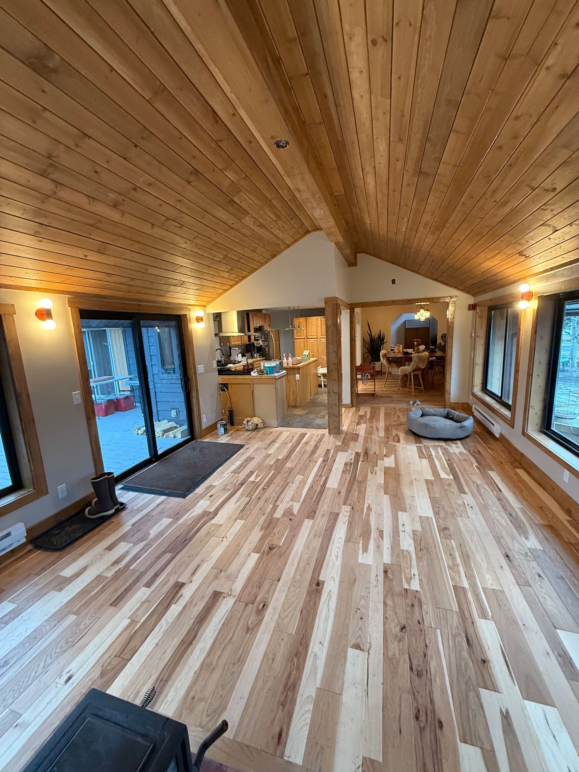 Wide wooden living room with vaulted plank ceiling, large windows, and a kitchen area in the back.