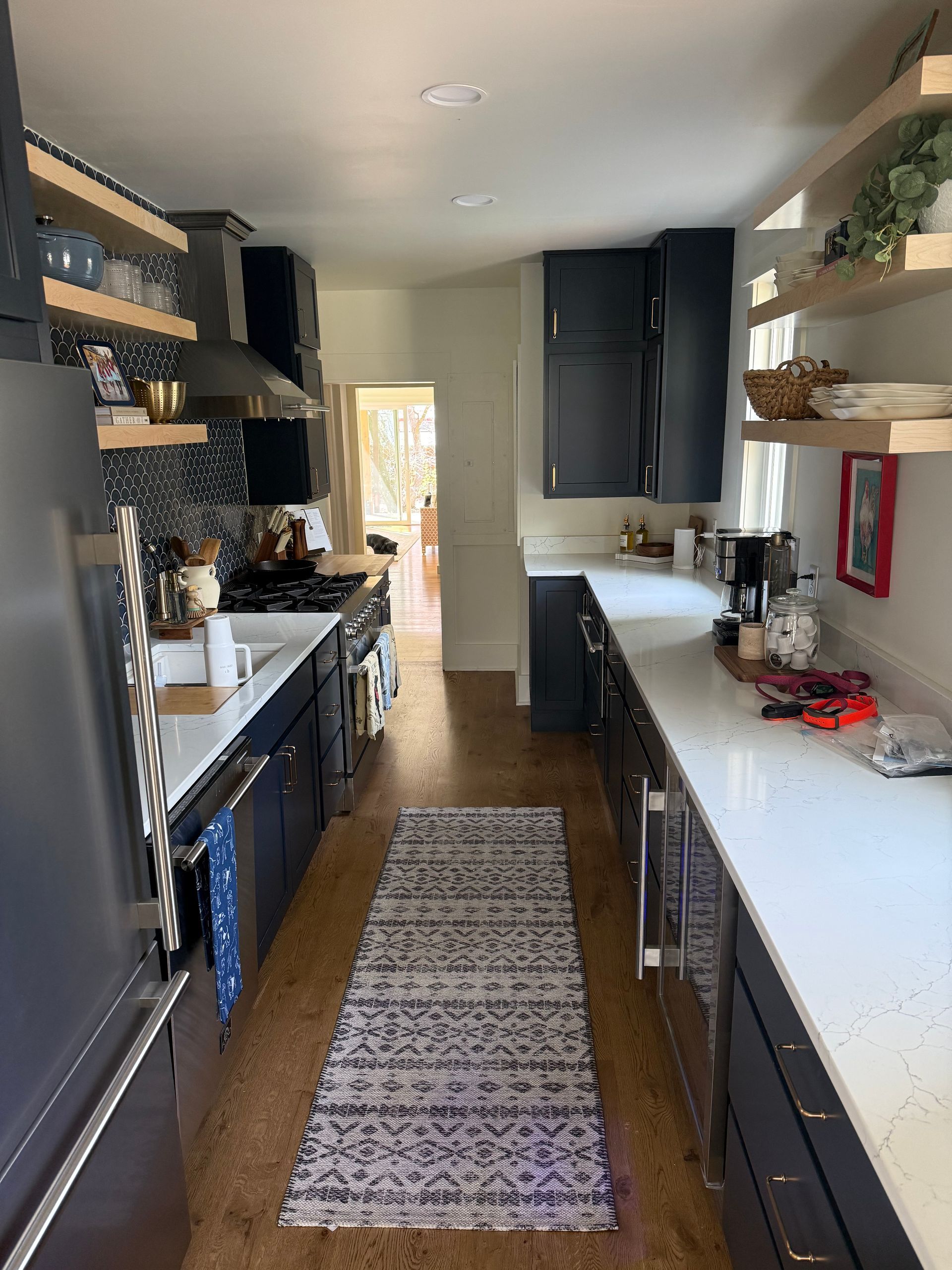 Narrow kitchen with dark cabinets, white counters, open shelves, and a patterned runner rug.