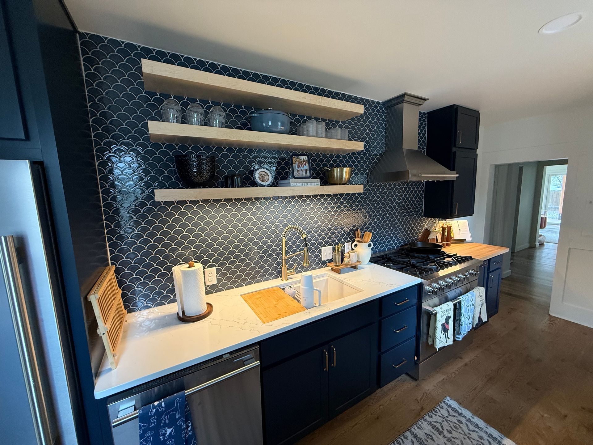 Modern kitchen with navy cabinets, white counters, patterned backsplash, open shelves, and stainless range hood.