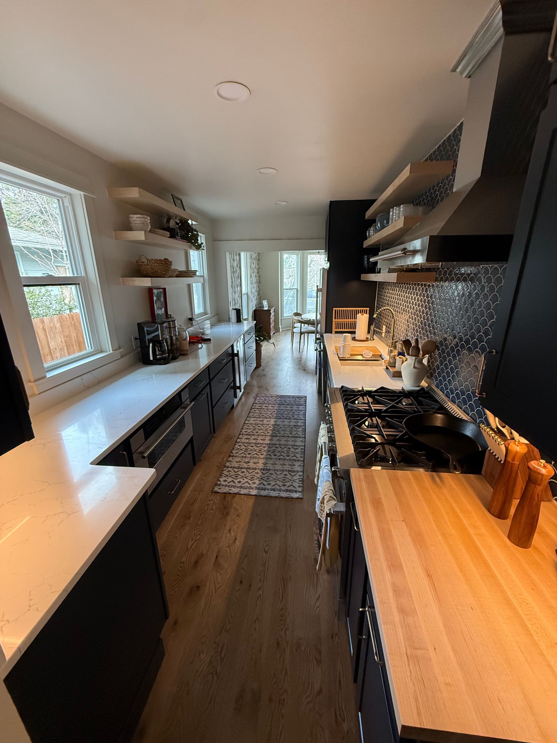 Narrow modern kitchen with white counters, dark cabinets, and a long window along the left wall.