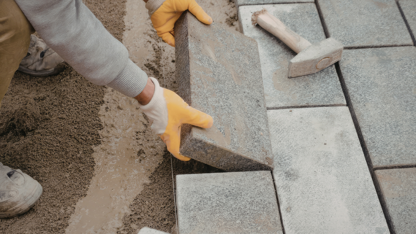 Hands in work gloves laying gray paving stones beside a bed of sand.
