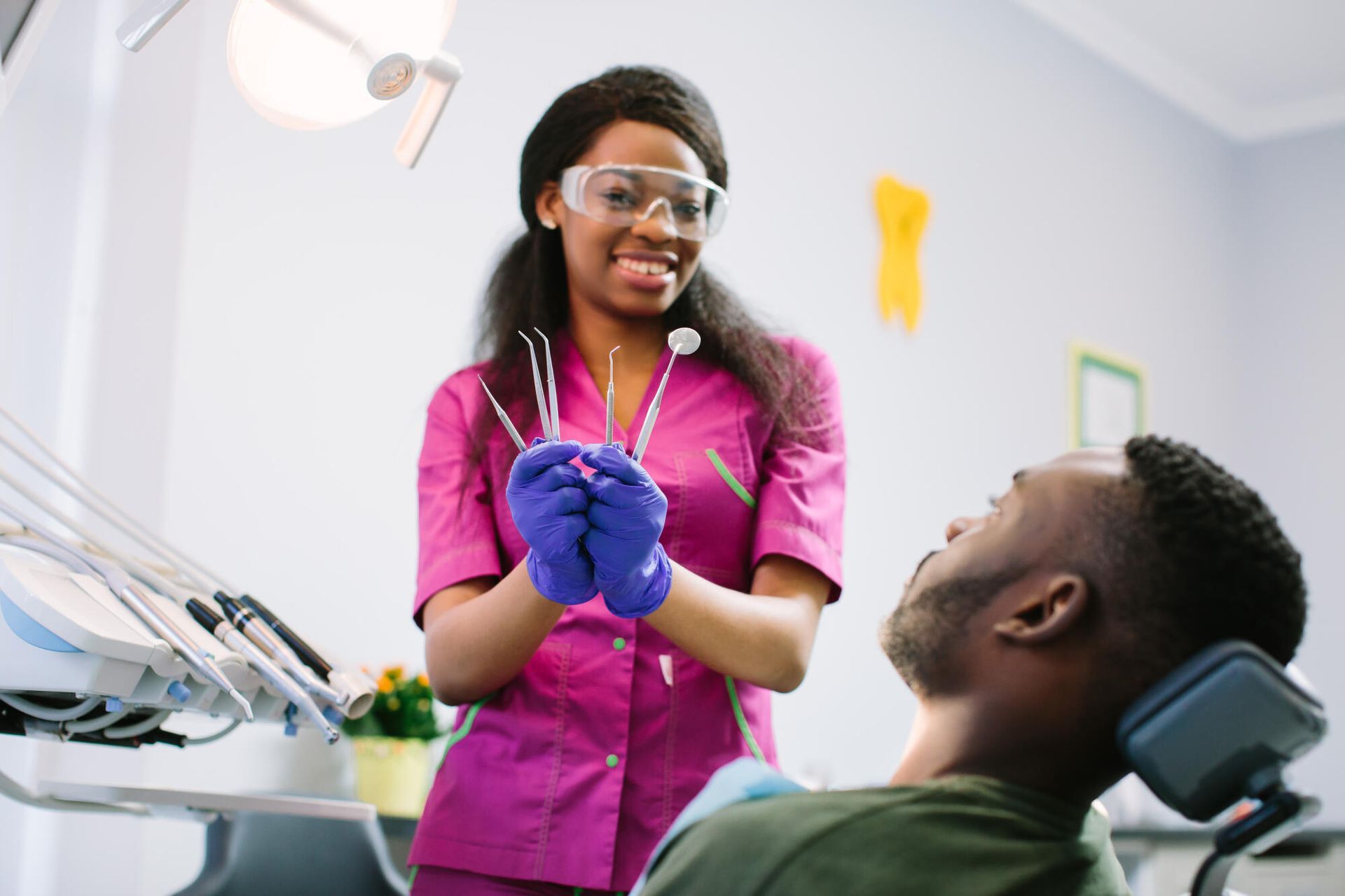 Dentist in pink scrubs adjusting gloves while speaking to a patient in a dental chair, with bright clinic equipment.