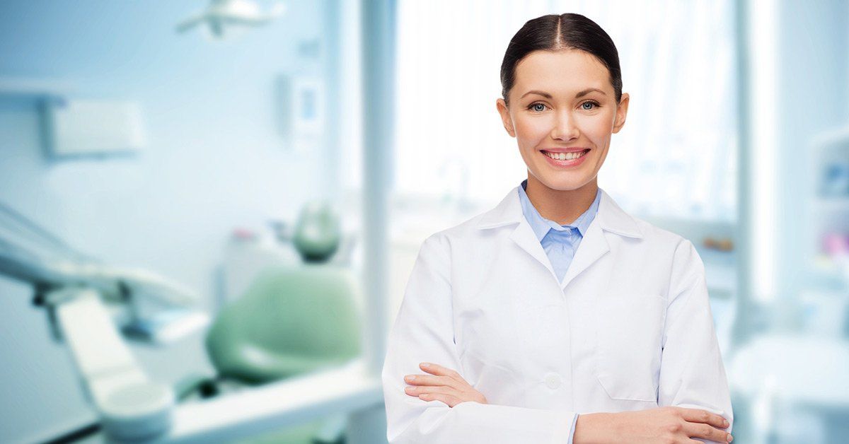 A female dentist is standing in a dental office with her arms crossed.